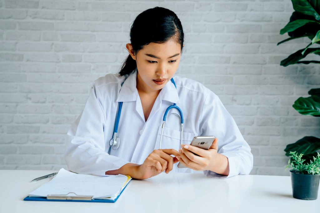 Young Asian female Doctor using a mobile phone at hospital. Healthcare worker scrolling and typing text messages at office