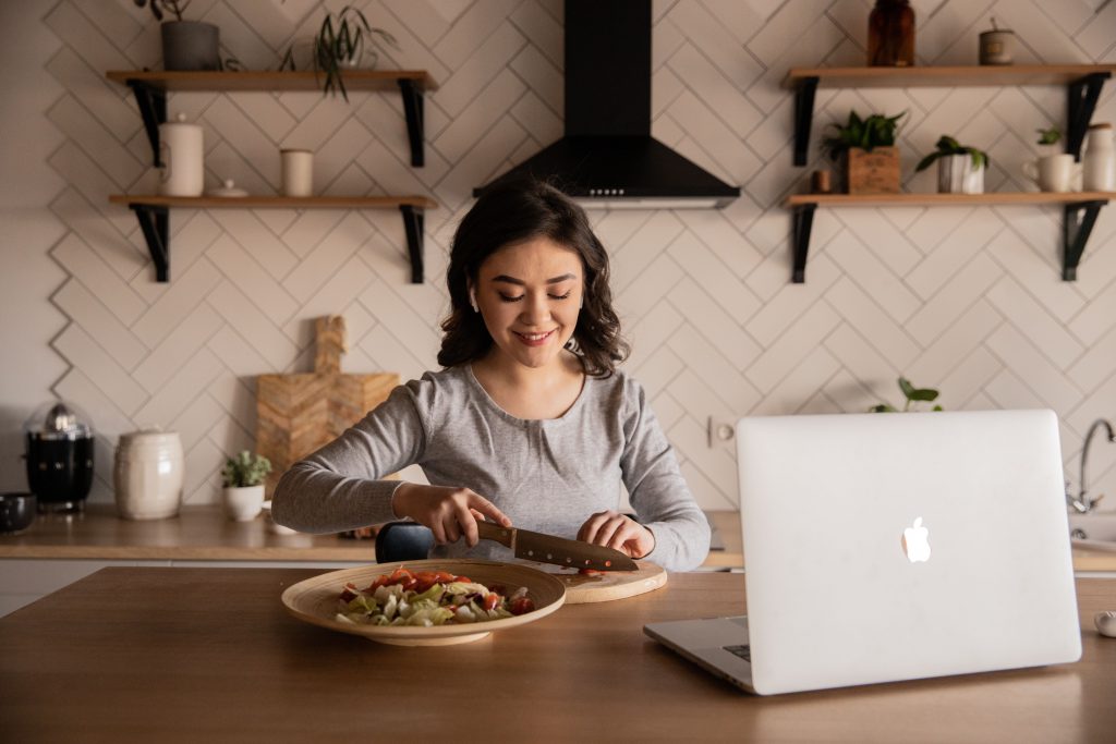 woman cuts vegetables while using a laptop.