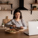 woman cuts vegetables while using a laptop.
