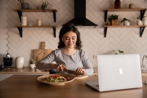 woman cuts vegetables while using a laptop.